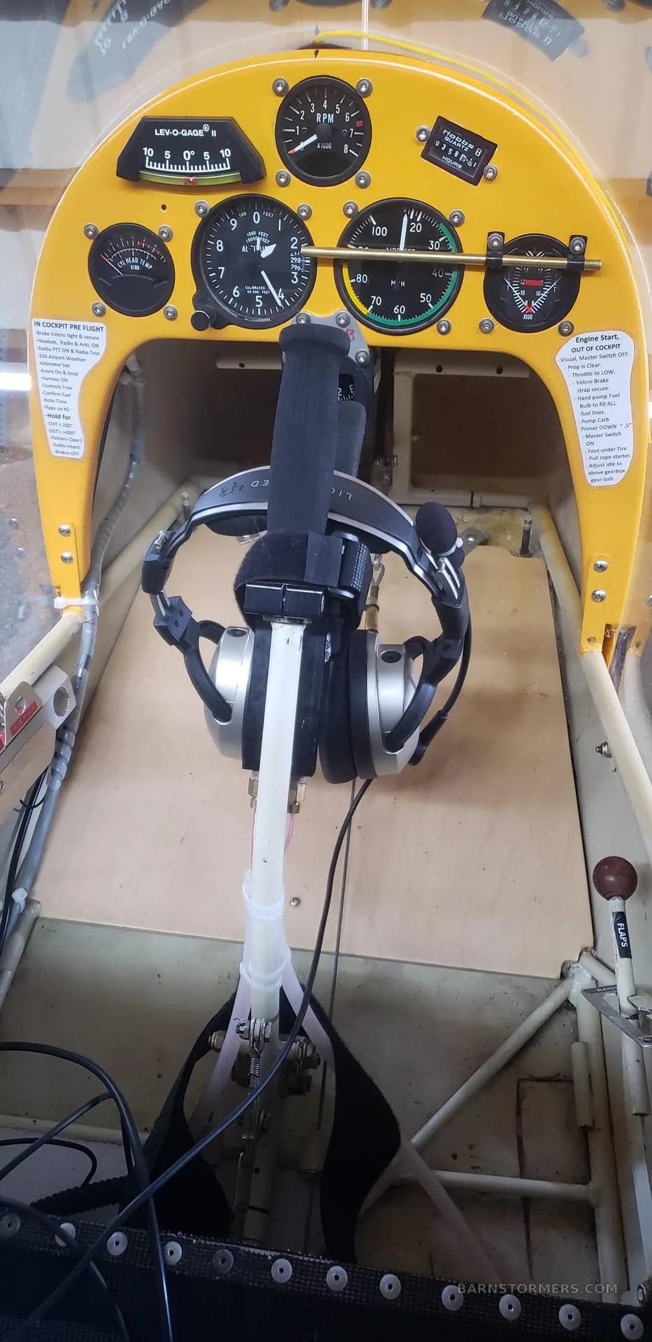 The yellow instrument panel of an ultralight aircraft shows gauges, a joystick, and a headset hanging from it.
