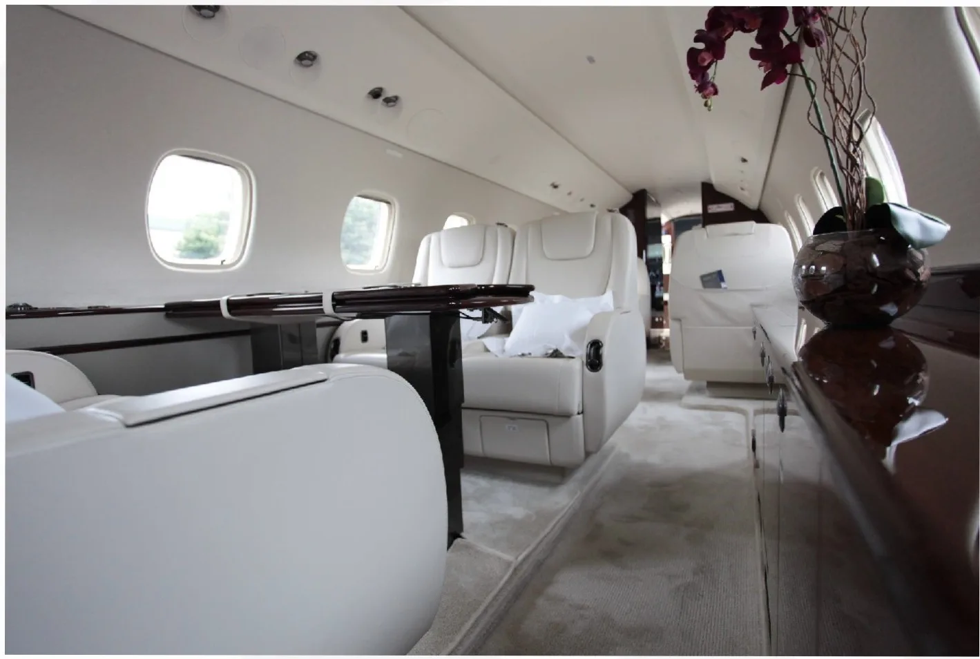 A wide-angle view of a private jet cabin, showing cream-colored seats, a dark wood table, and a vase with purple flowers.