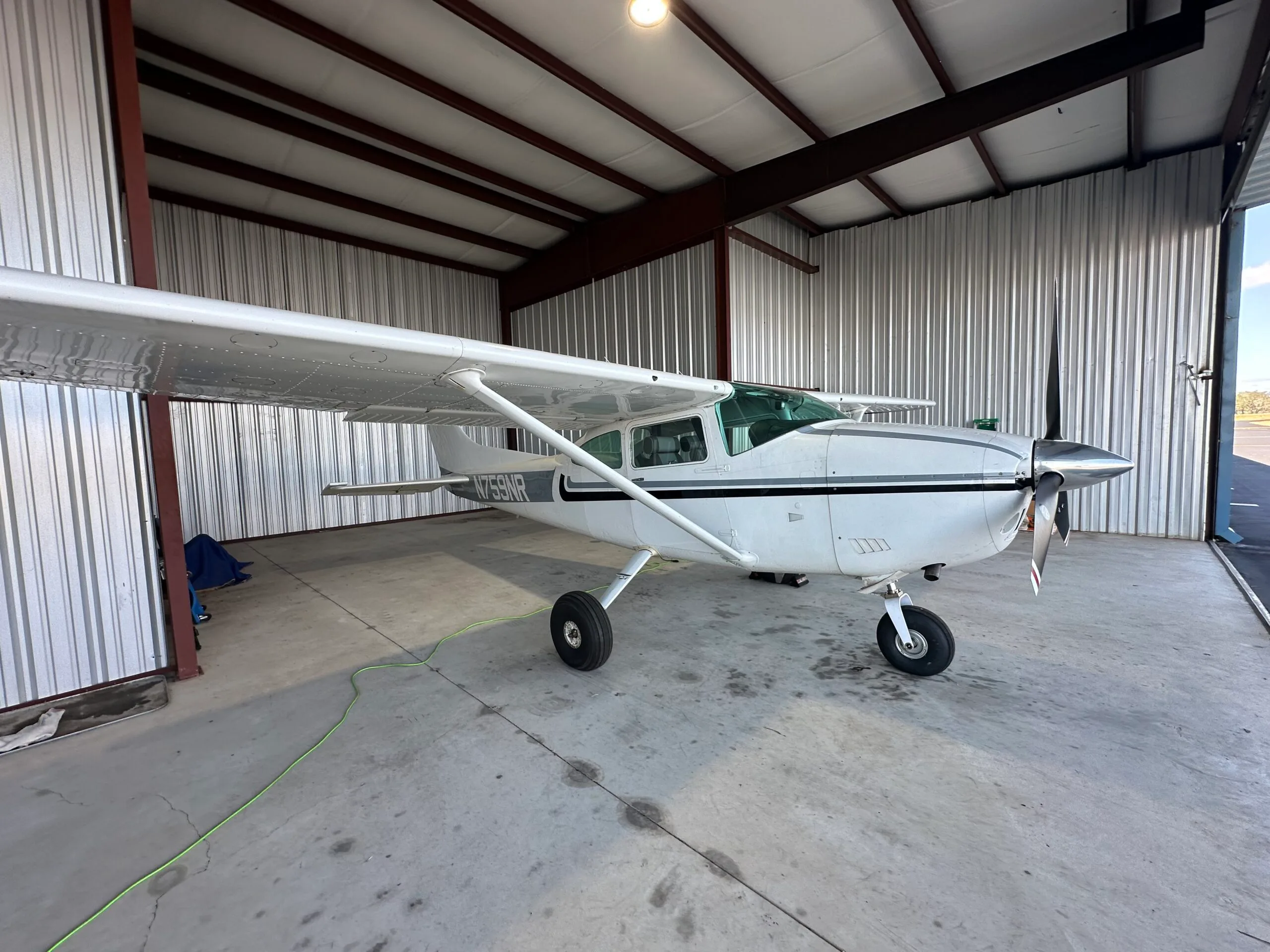 A white single-engine propeller aircraft with black and grey stripes, registration N759NR, parked inside a metal hangar.