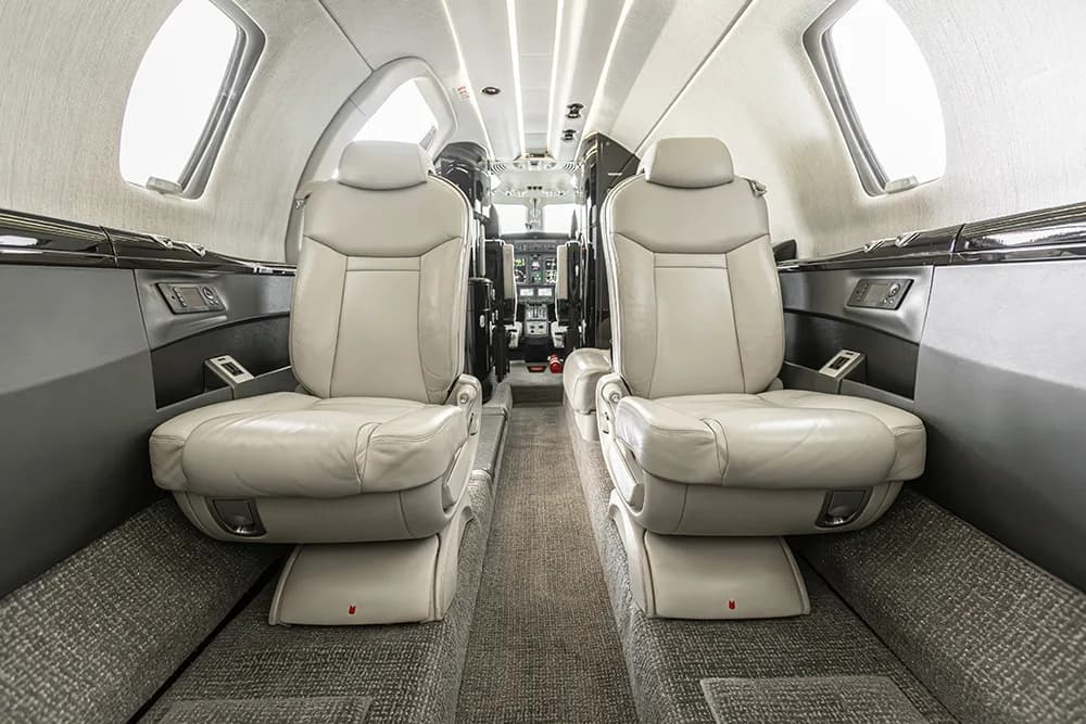 A view inside the private jet cabin, showing two rows of light grey seats facing forward towards the cockpit.