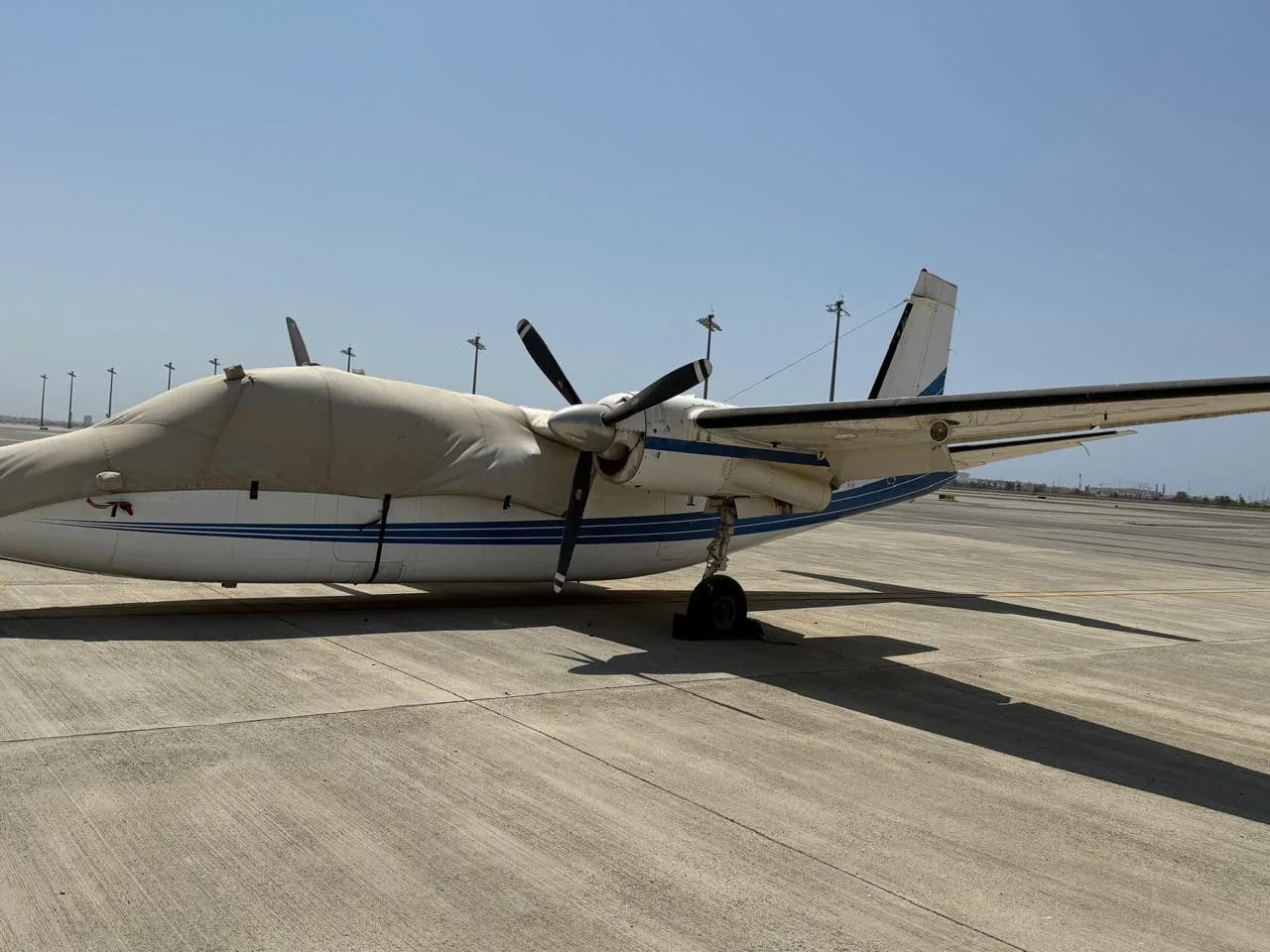 A white and blue twin-engine turboprop aircraft is parked on an airfield, with its nose covered and propellers visible.