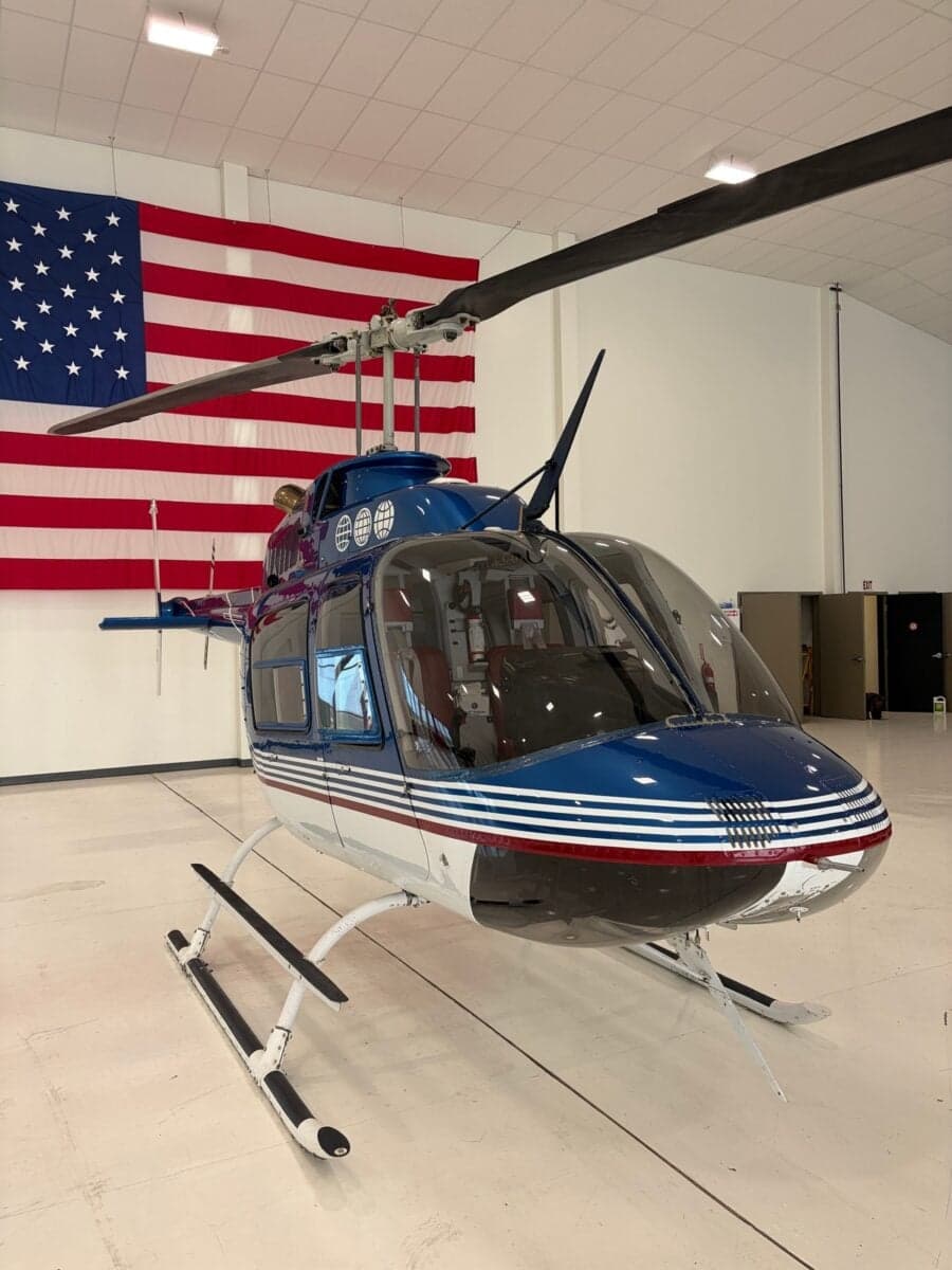 A blue and white Bell 206B JetRanger helicopter parked in a hangar with an American flag in the background.