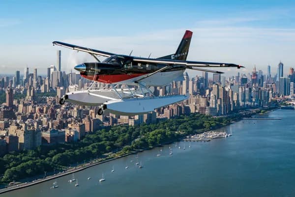 A black and red Quest Kodiak 100 seaplane flying over a city skyline and river.