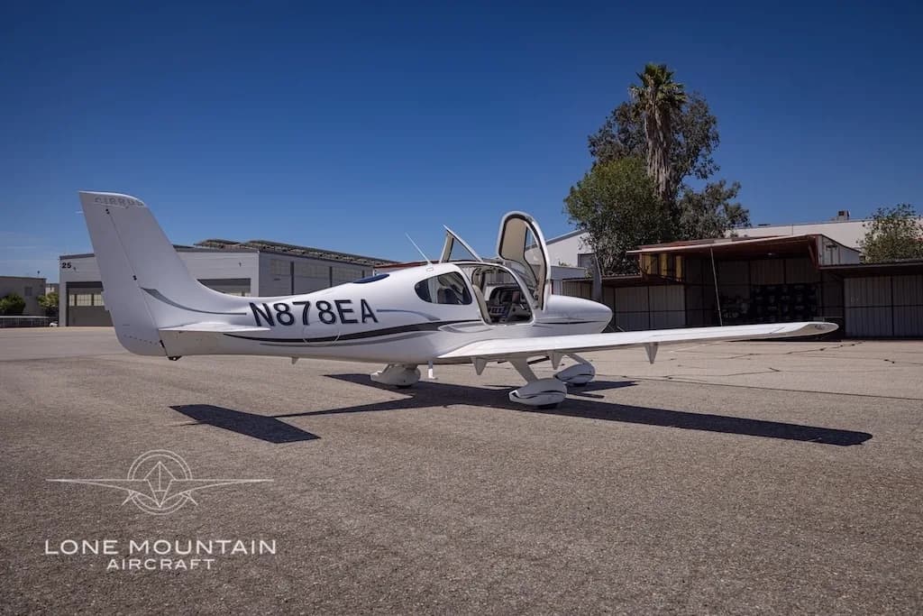 A white Cirrus aircraft with registration N878EA and open doors is parked on an airfield next to buildings and a palm tree.