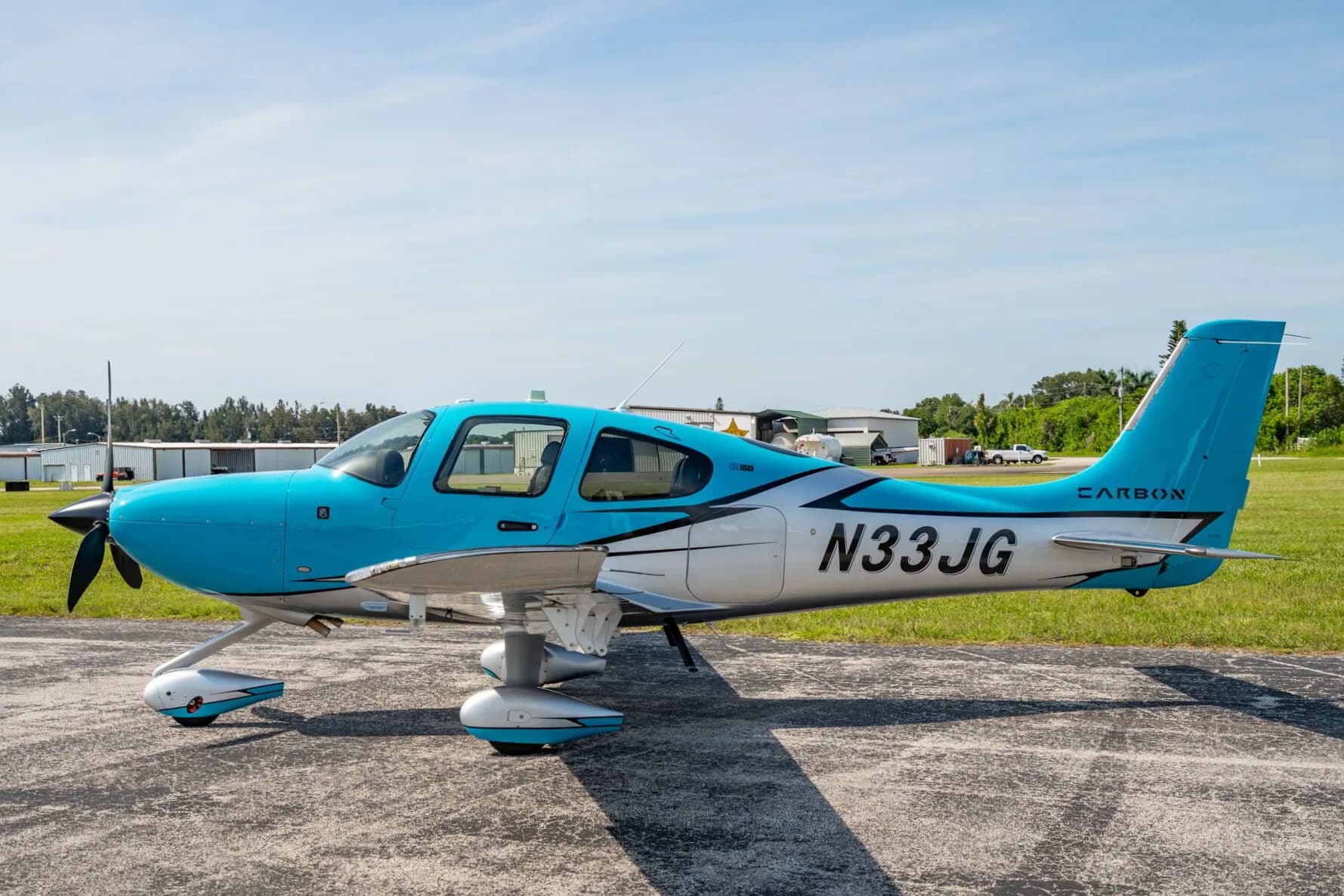 A side view of a blue and silver Carbon aircraft (N33JG) parked on an asphalt ramp next to a grassy field.