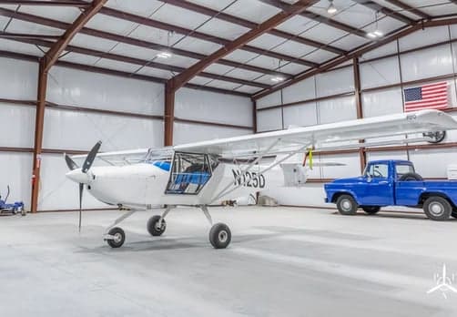 A white light sport aircraft, registration N125D, is parked in a hangar next to a blue pickup truck.