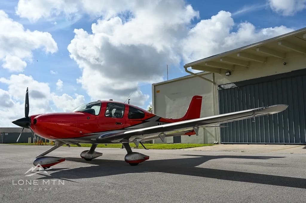 A side view of a red and silver Cirrus SR22 aircraft, registration N273RV, parked on an asphalt ramp next to a hangar.