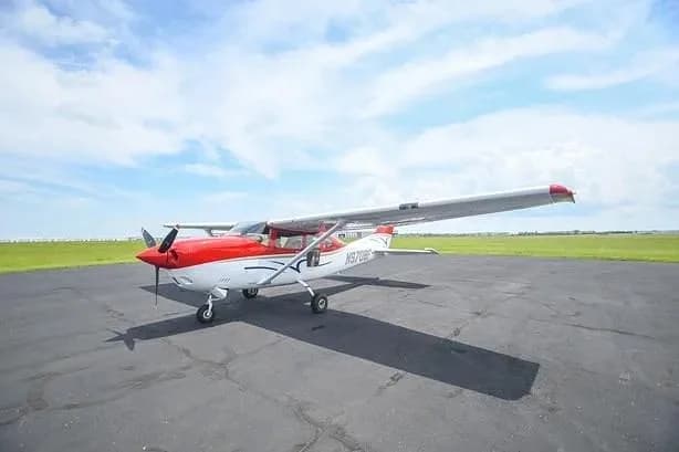 A red and white single-engine propeller aircraft (Cessna 172) parked on an asphalt runway under a partly cloudy sky.