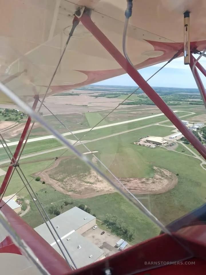 Aerial view from a red biplane, showing its wing and struts over an airfield and surrounding rural landscape.