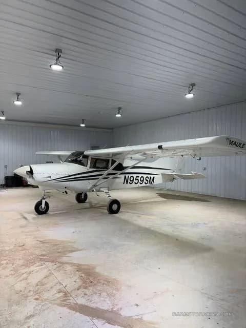 A white Maule M-7-235B aircraft with black stripes and registration N959SM is parked inside a hangar, viewed from the front-left.