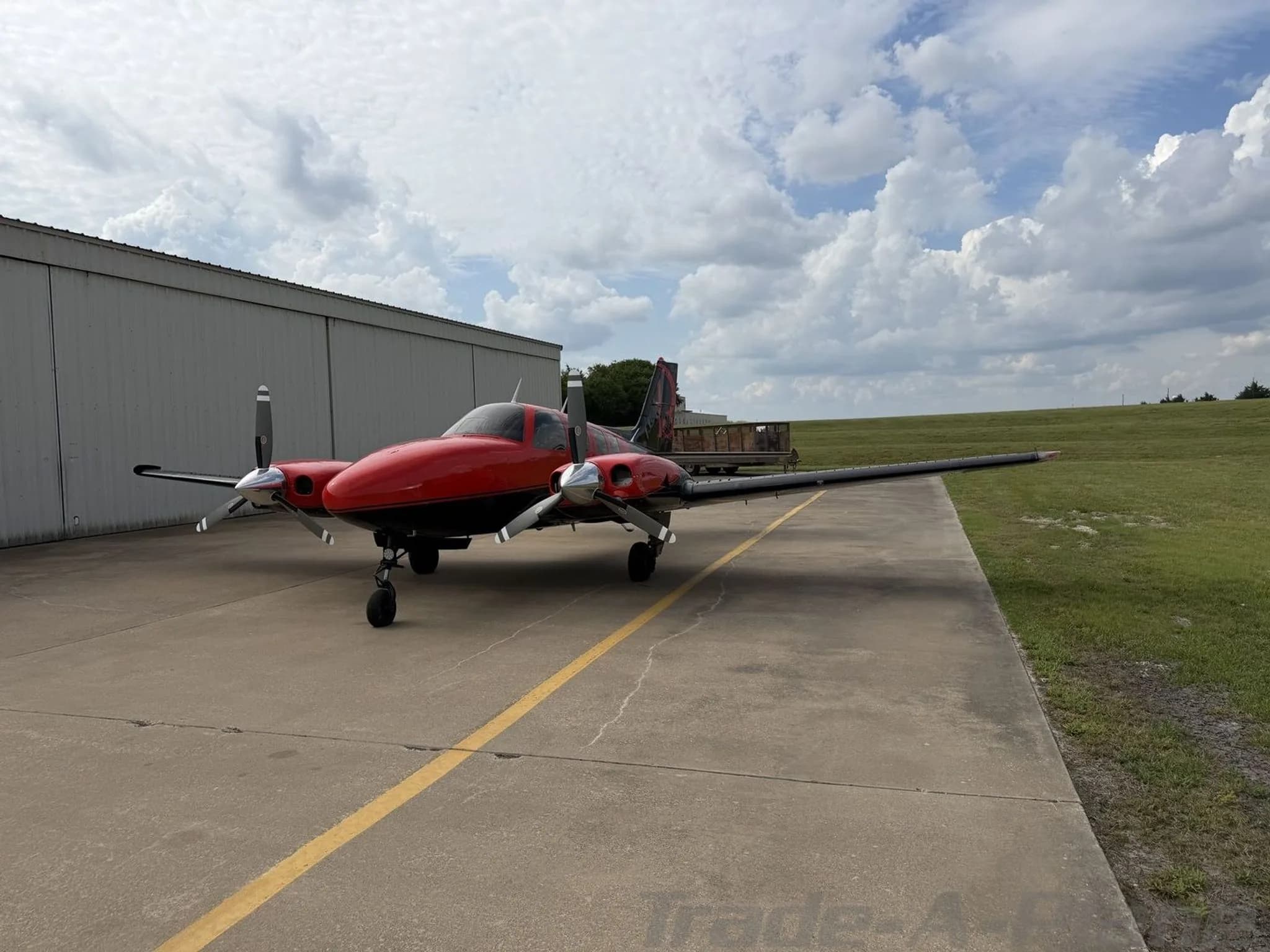 A red and black twin-engine propeller aircraft, N10NF, parked on an airfield tarmac next to a hangar.