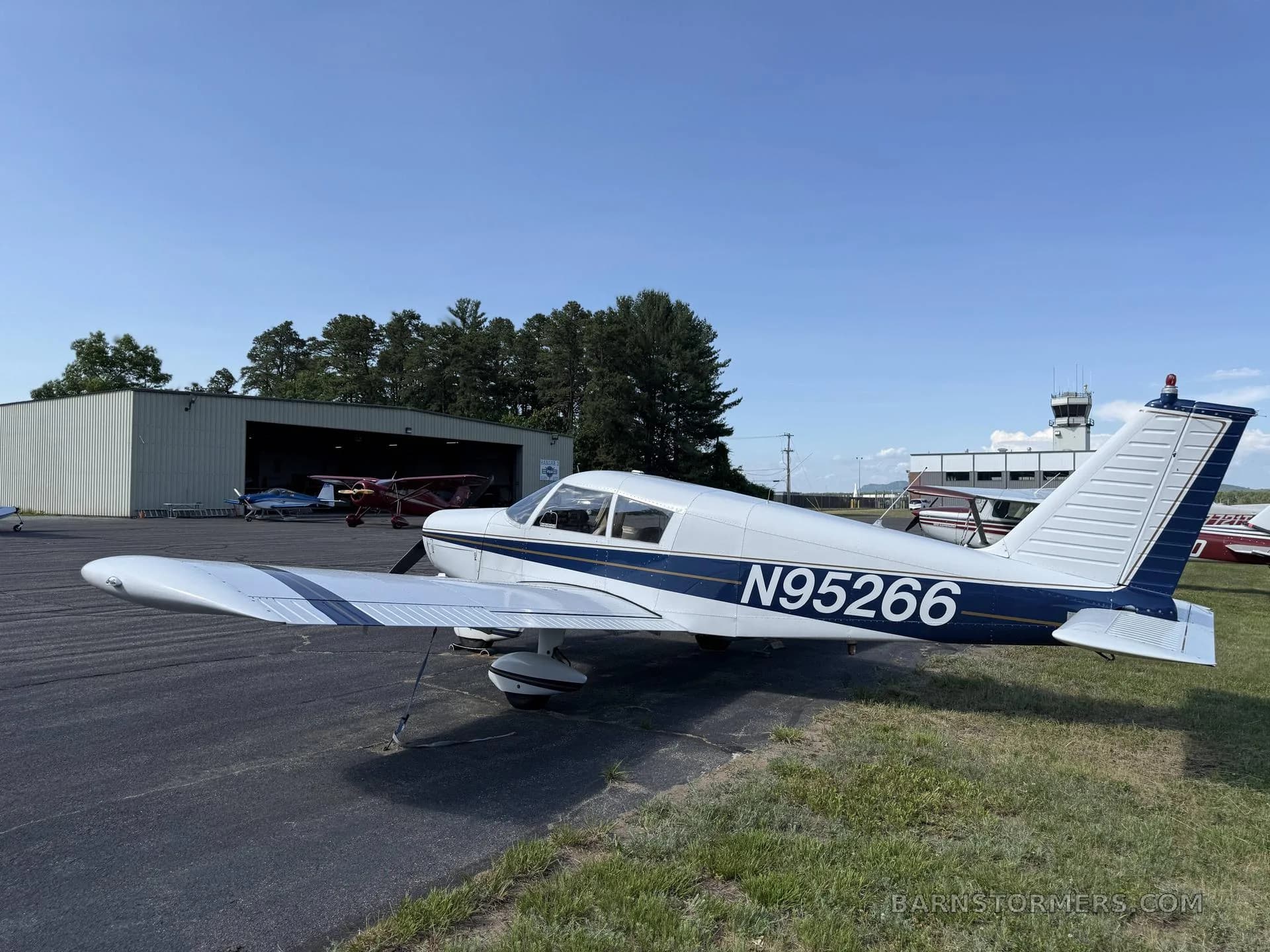 A white single-engine propeller aircraft (N95266) with blue stripes, parked on an airport apron with an open hangar and control tower in the background.