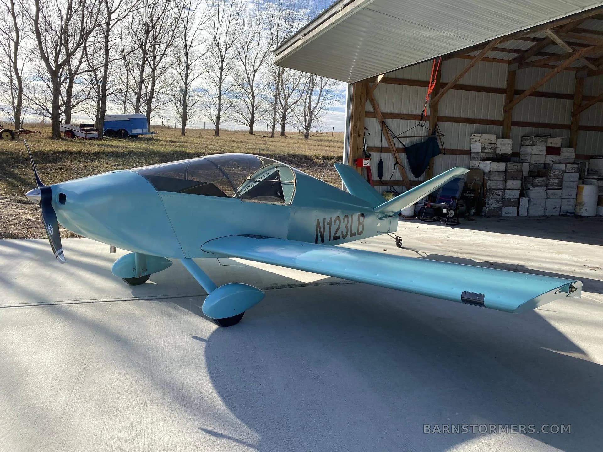 A light blue single-engine experimental aircraft (N123LB) with a bubble canopy, parked on concrete outside a hangar with trees in the background.