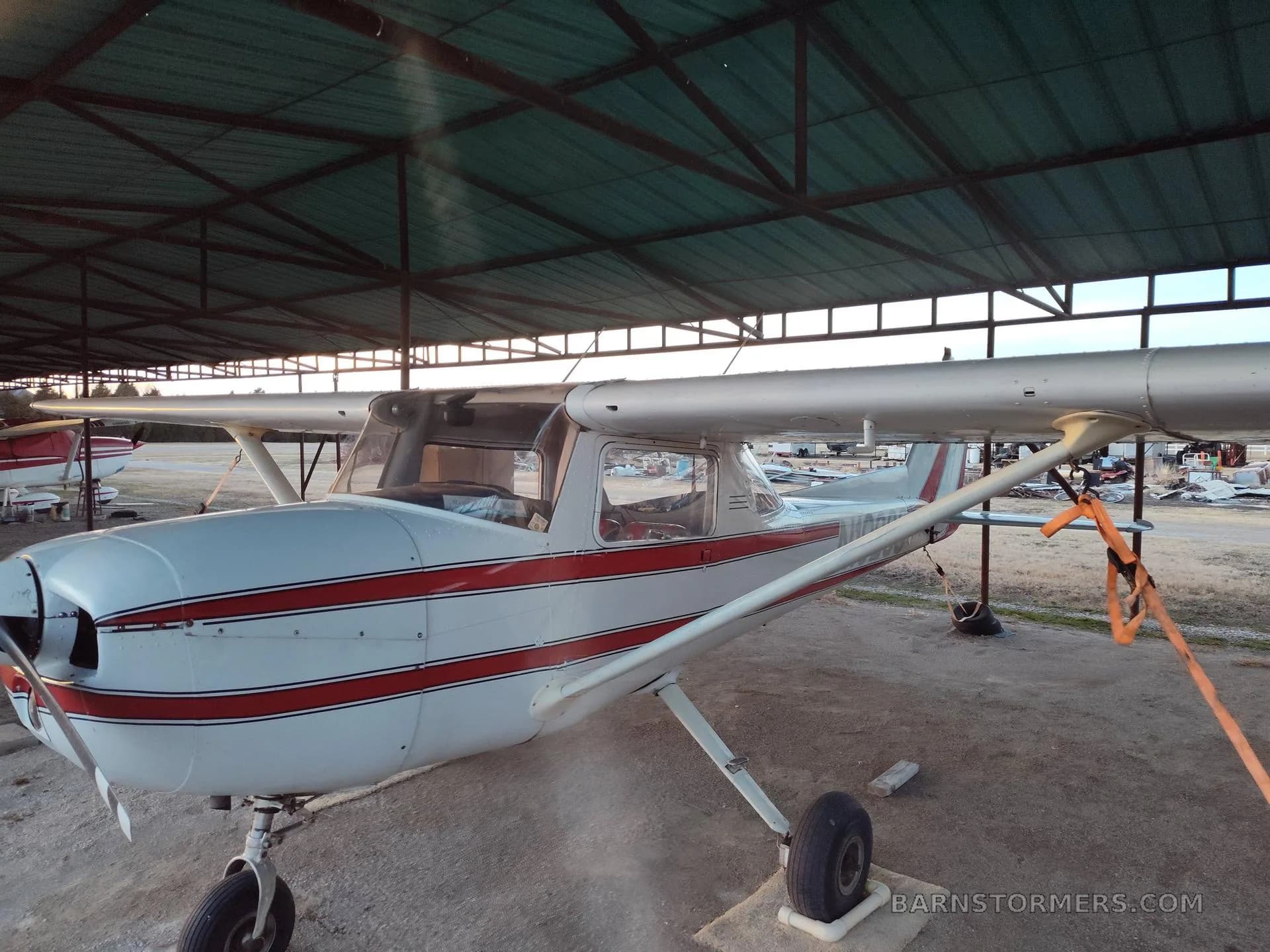 A white and red striped Cessna 150/152 type aircraft is parked under a metal hangar roof.