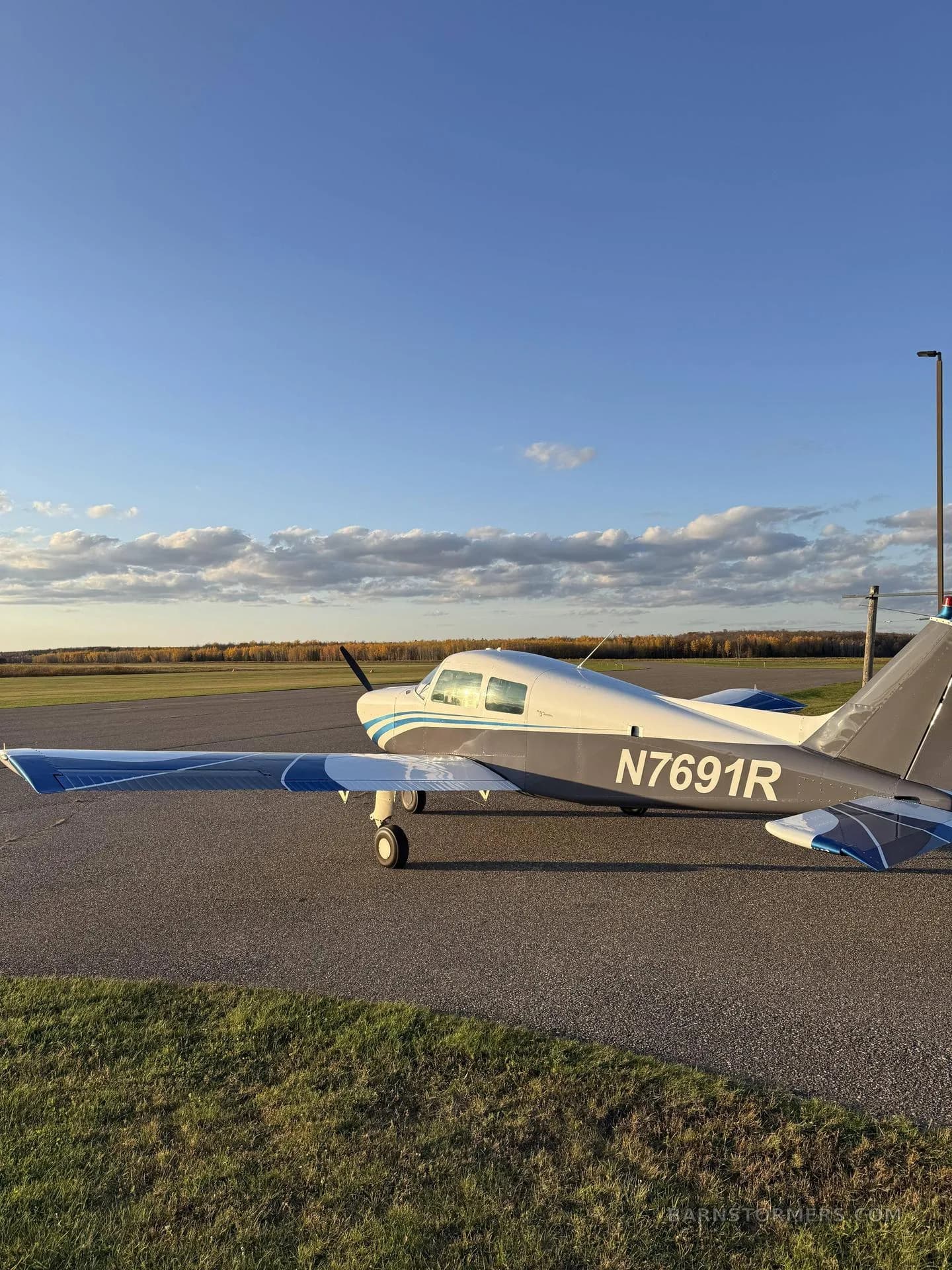 A white and blue single-engine aircraft (N7691R) parked on an asphalt ramp with a grassy field and distant trees under a partly cloudy sky.