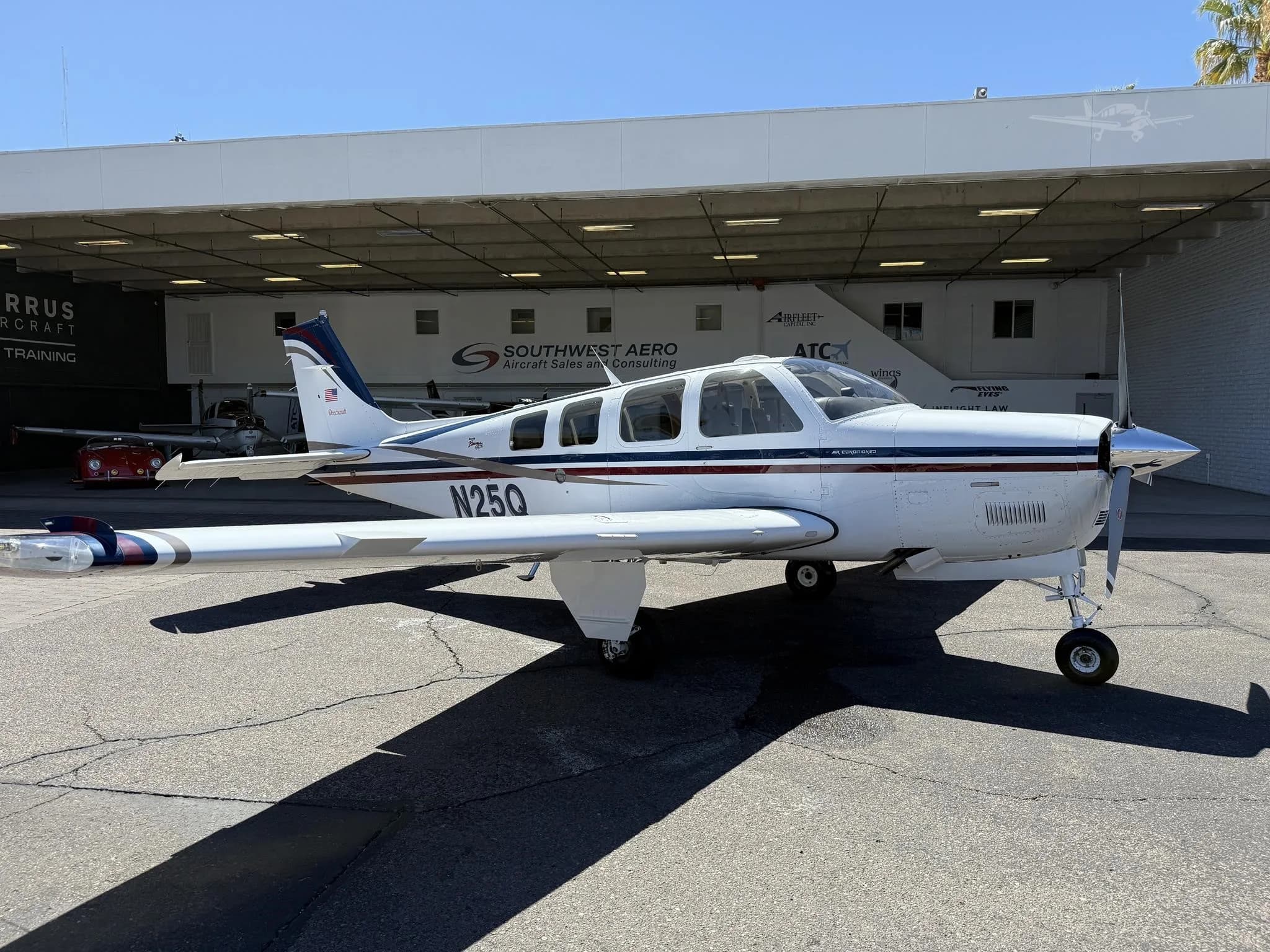 A white Beechcraft Bonanza with blue and red stripes is parked on a tarmac in front of a hangar.