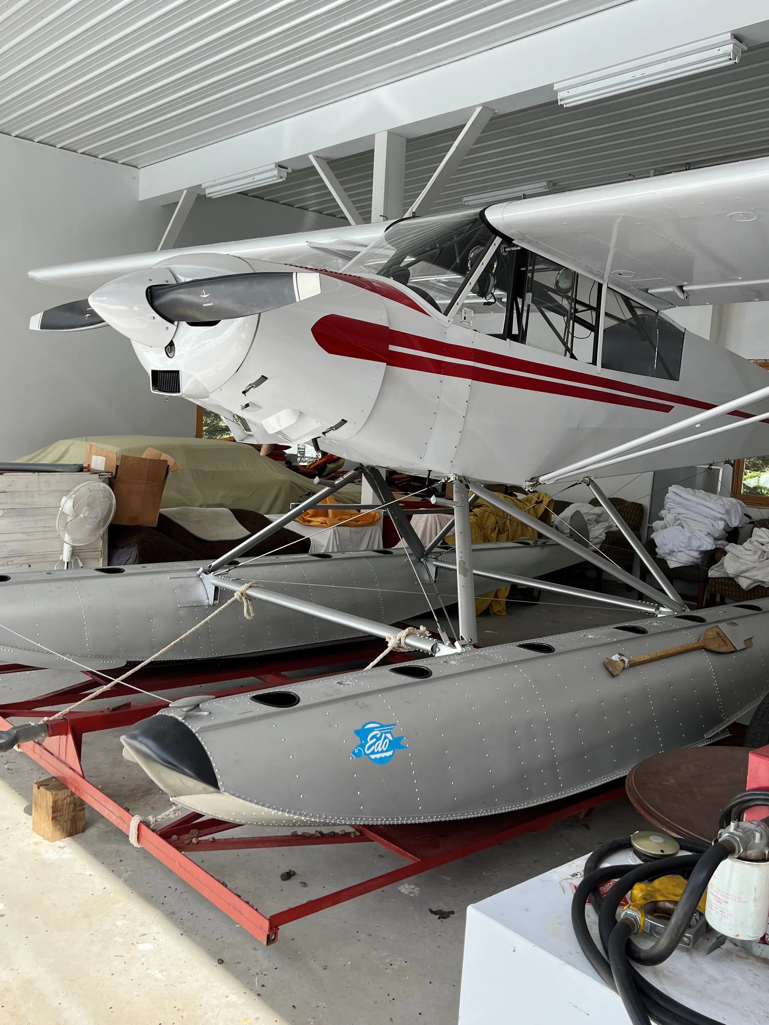 A white floatplane with red stripes is parked indoors on a red dolly, viewed from the front-right, showing its Edo floats and a paddle.