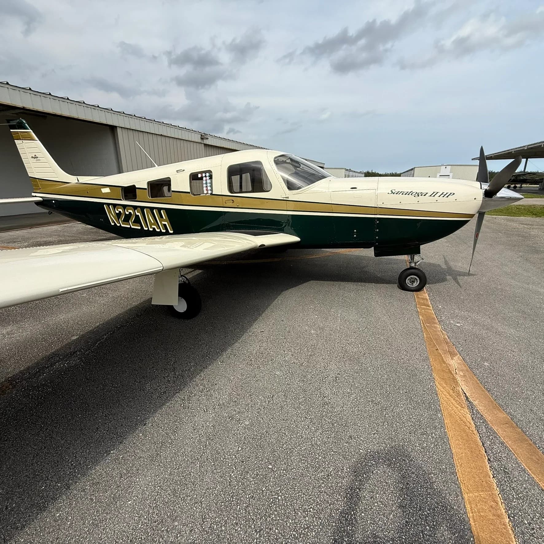 Side view of a Piper Saratoga II HP, N221AH, with a white, gold, and dark green paint scheme, parked on an airport tarmac.