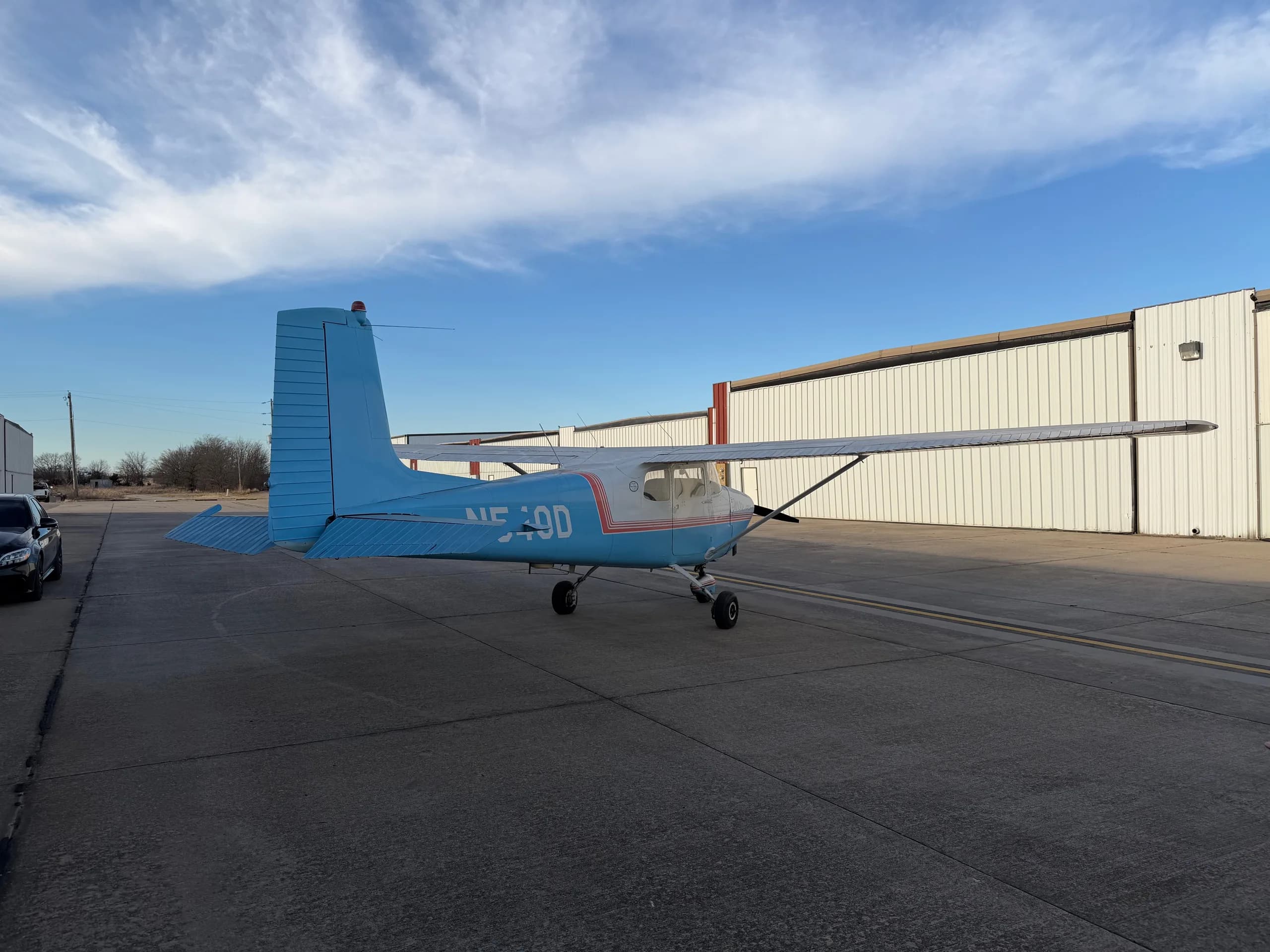 A rear-left view of a white and light blue Cessna 172, registration N540D, parked on an airfield with hangars in the background.