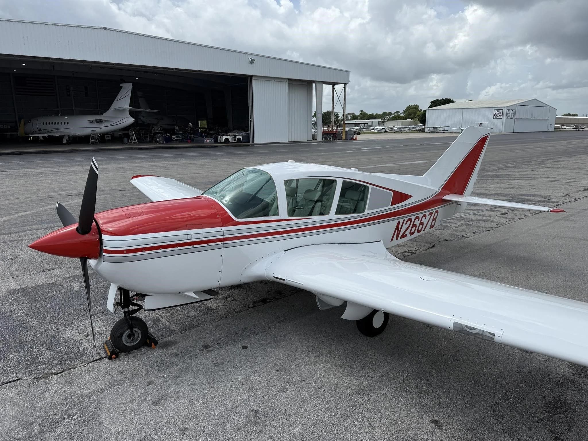 A white and red single-engine aircraft, N2667B, parked on an airport tarmac with hangars in the background.