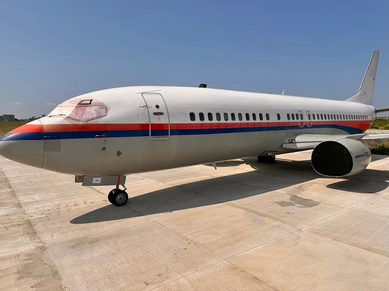 A large white passenger jet with red and blue stripes is parked on a concrete tarmac under a clear sky.