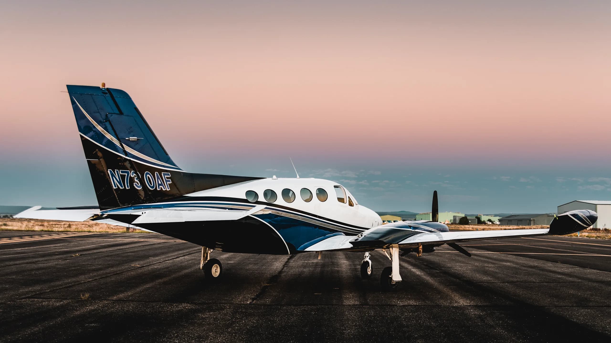 A white and blue twin-engine aircraft with propellers visible, parked on a runway at sunset.