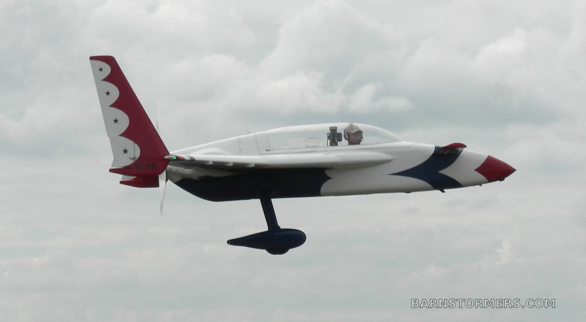 A white, red, and blue aircraft with twin tails and a pusher propeller, flying against a cloudy sky, with a pilot visible in the cockpit.