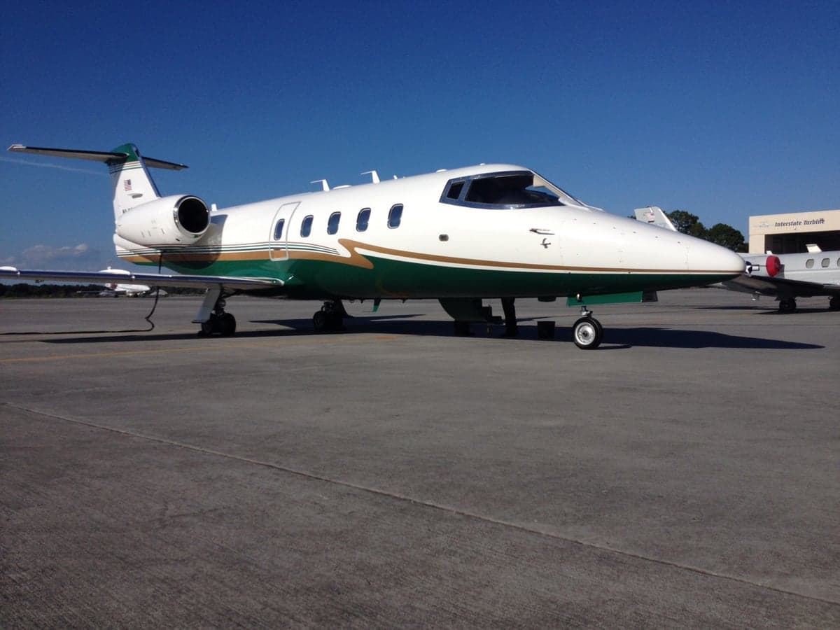 A white, green, and gold Learjet is parked on an airport tarmac under a clear blue sky.