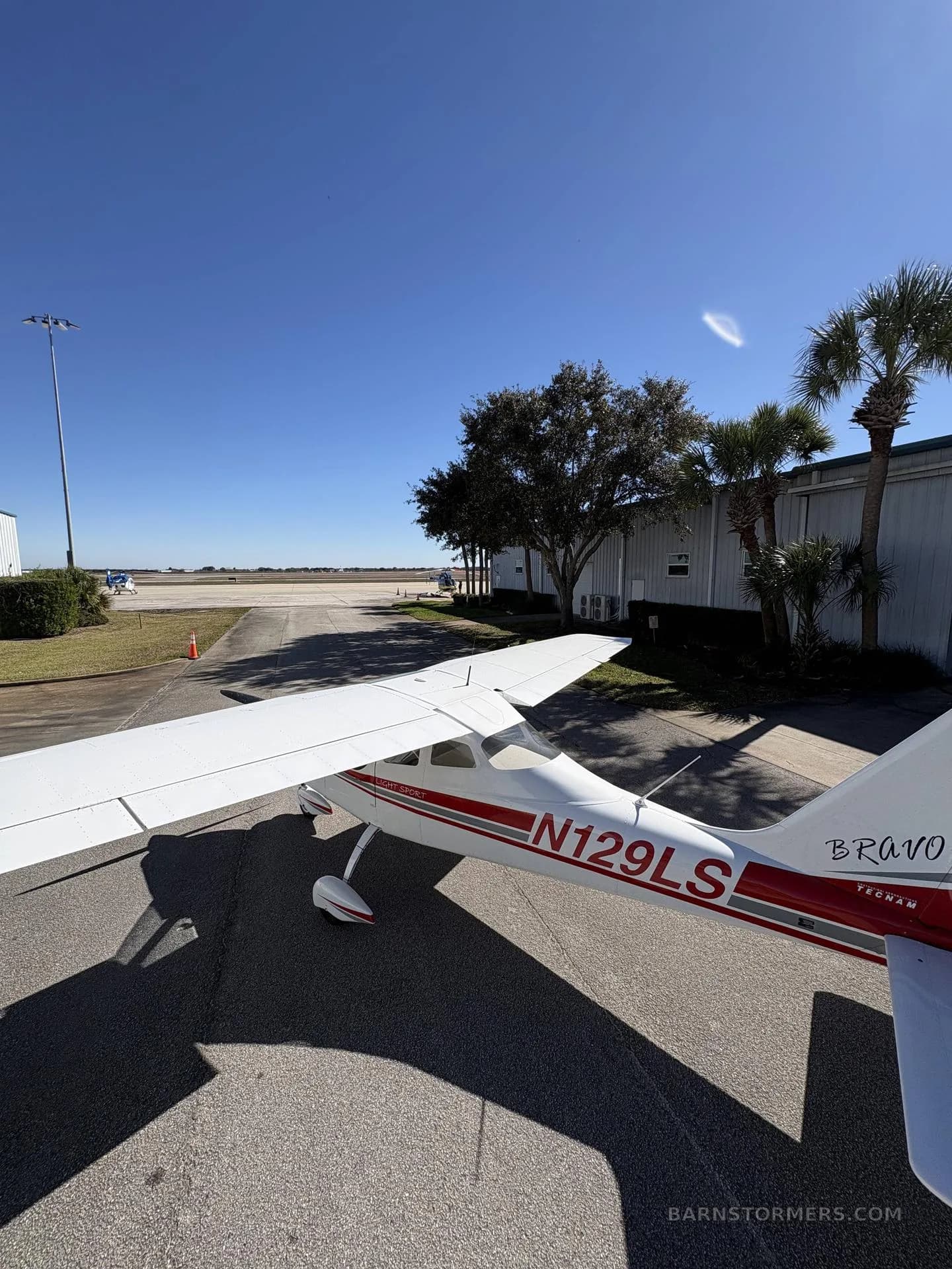 A white and red Tecnam P2008 aircraft with registration N129LS is seen from the rear-right, parked on an asphalt runway with palm trees and hangars in the background.