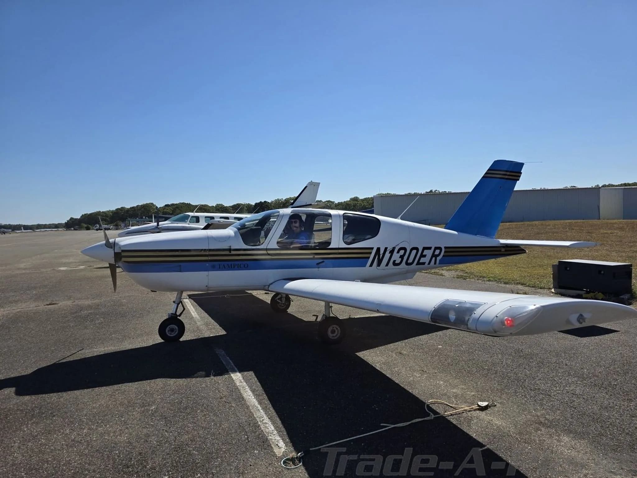 A white and blue Socata TB-9 Tampico aircraft with tail number N130ER parked on an asphalt ramp on a sunny day.