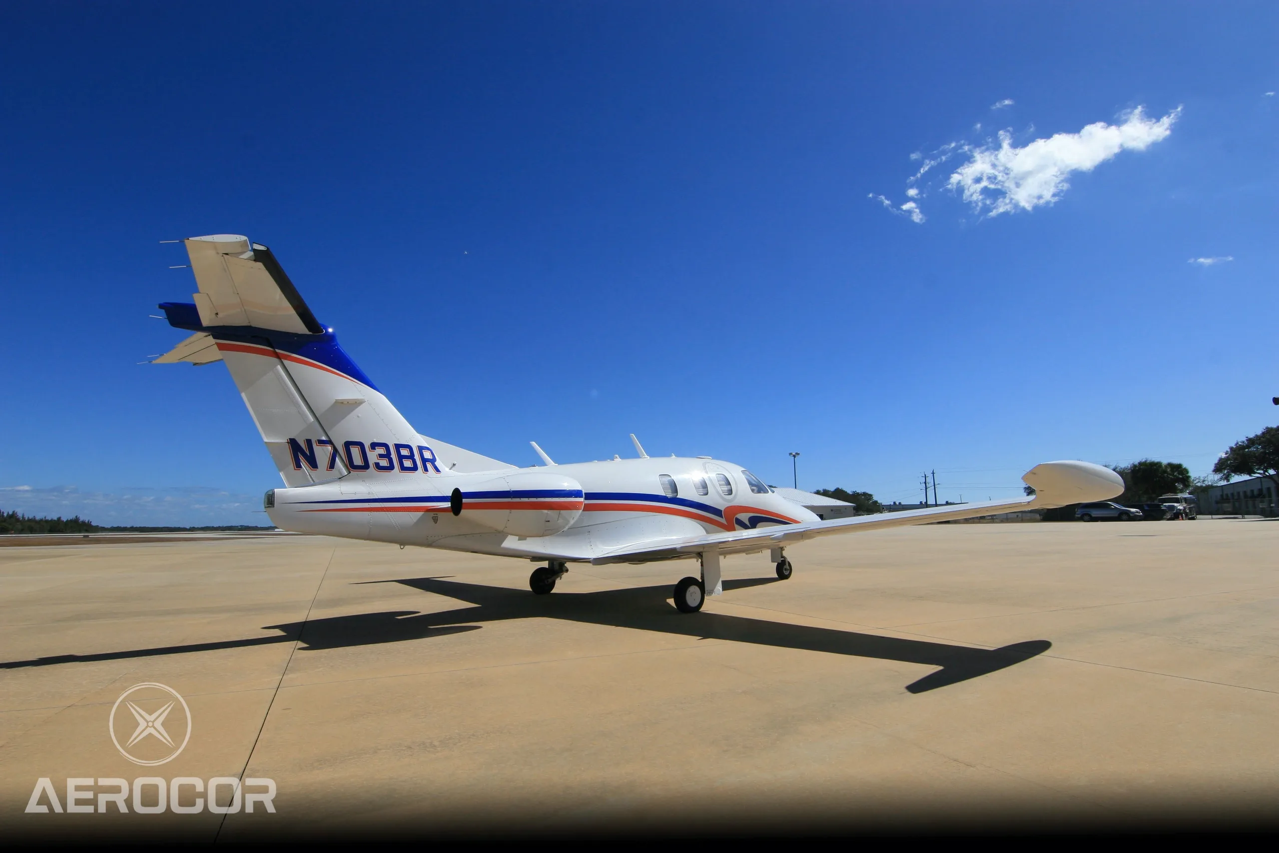 A rear-side view of a white Eclipse 500 jet with blue and orange stripes, tail number N703BR, parked on an airfield.