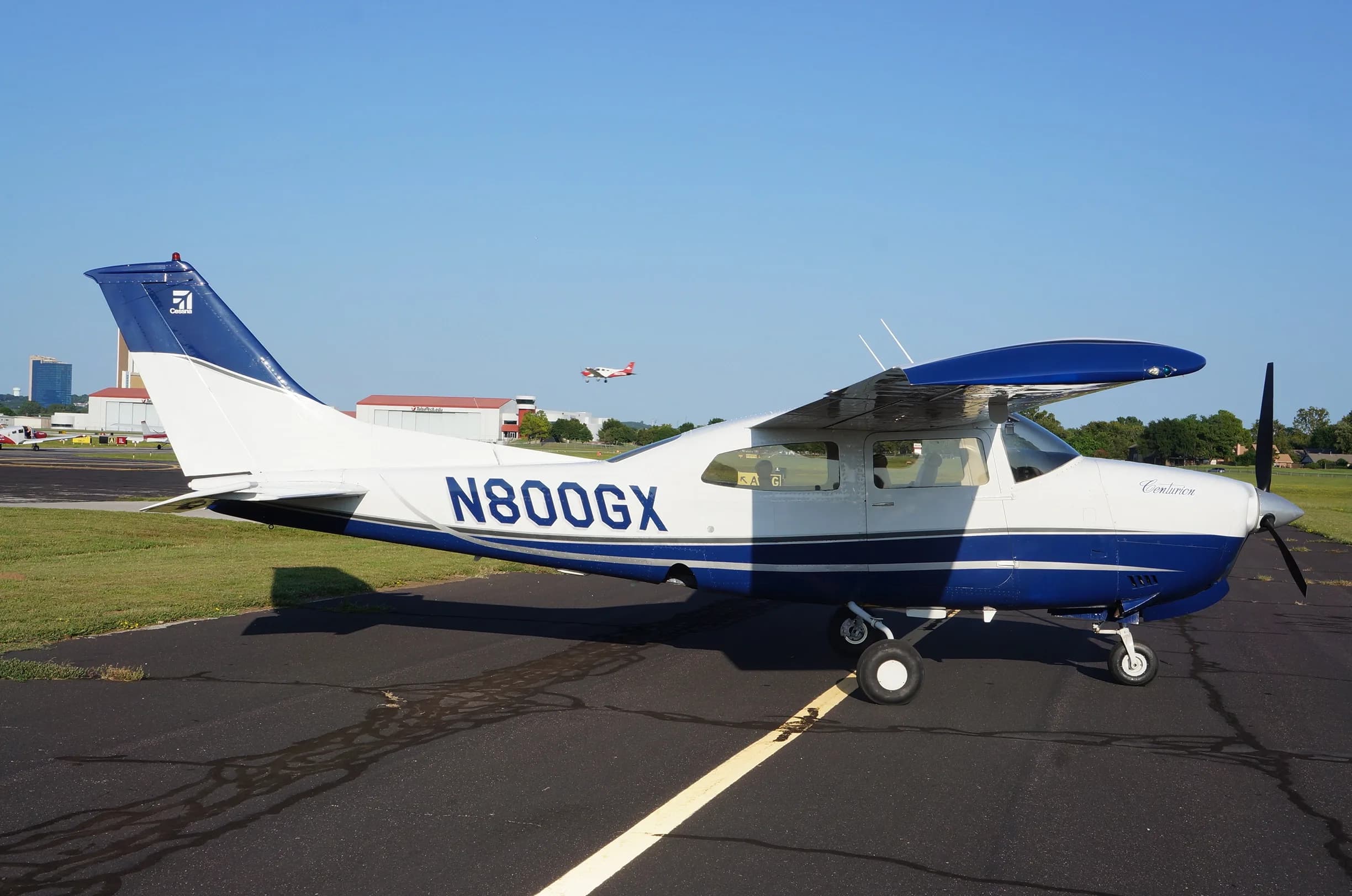 A white and blue Cessna 206 Stationair aircraft, N800GX, is parked on an airport tarmac with buildings and another plane in the distance.