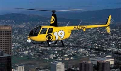 Yellow Robinson R44 helicopter with black tail in flight over a city.