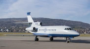 A white and blue Dassault Falcon business jet, tail number N121B, is parked on an airport tarmac with mountains in the background.