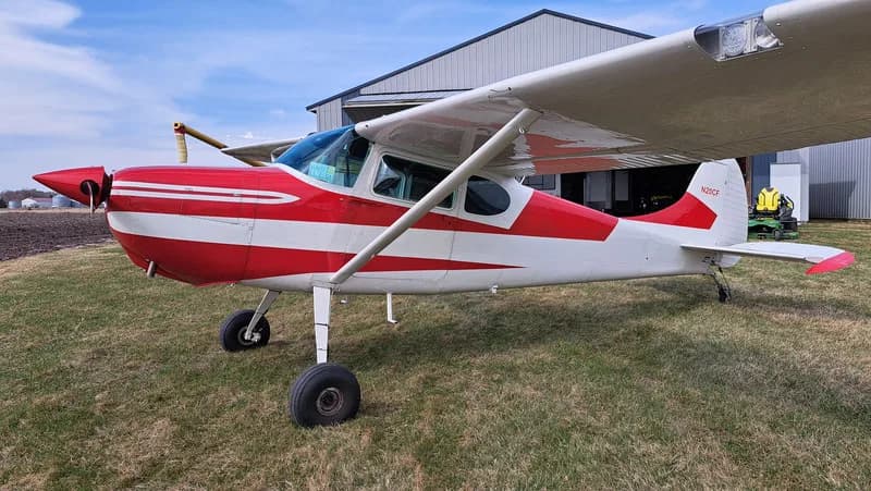Side view of a red and white Cessna on a grassy field in front of a hangar.