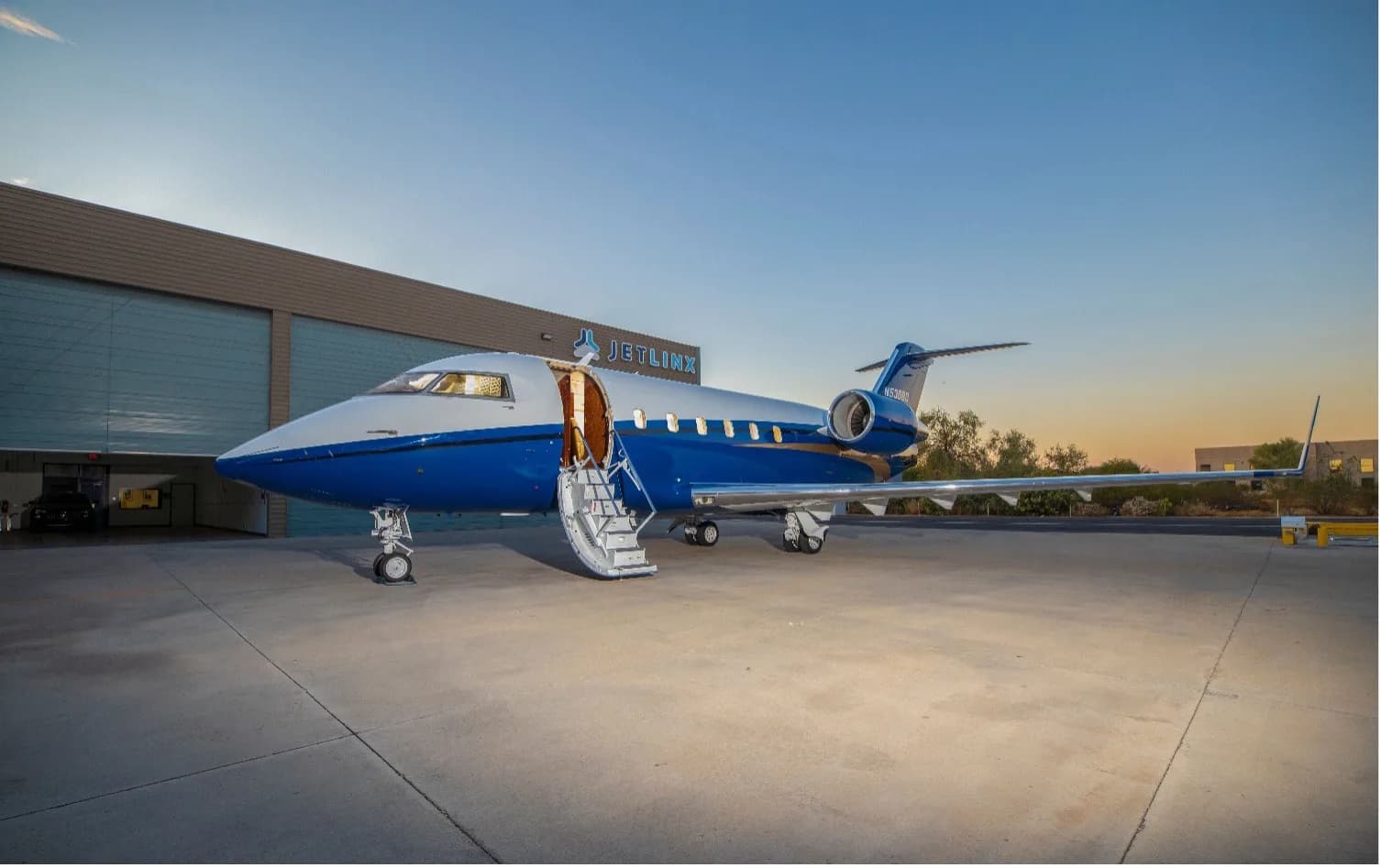 A Bombardier Challenger 604 business jet with a blue and white paint scheme, registration N530BD, parked on a tarmac with its door open at sunset.