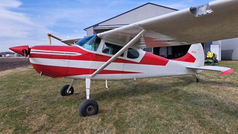 A side view of the red and white Cessna on a grassy field in front of a hangar.
