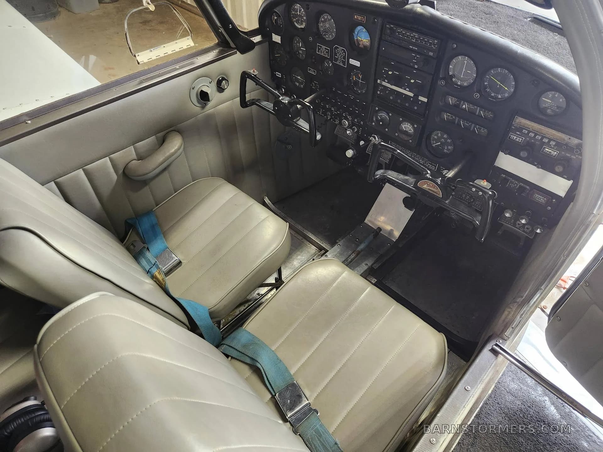 The light gray interior cabin and cockpit of a Piper Cherokee aircraft, showing the front seats with blue seatbelts.