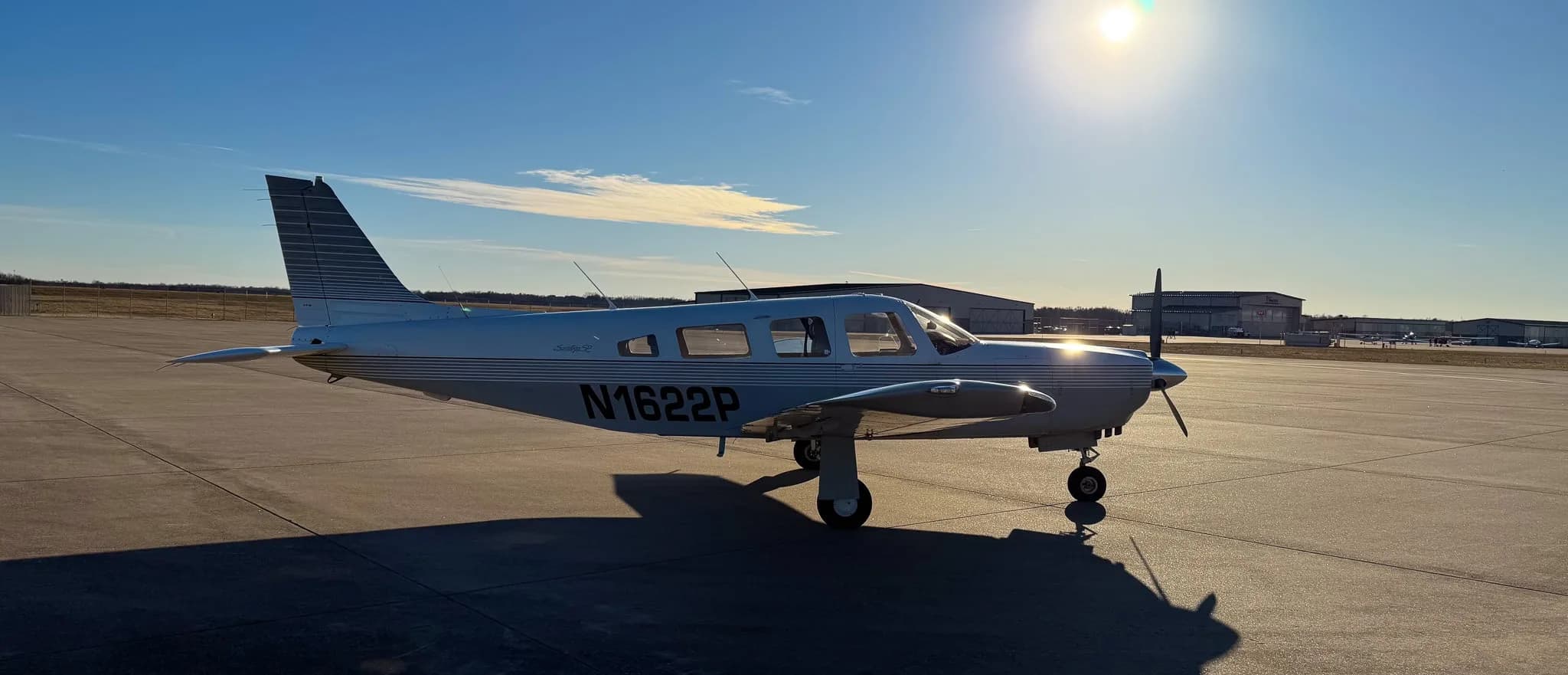 A side view of a white Piper Saratoga SP (N1622P) with grey stripes on an airfield with the sun low in the sky.