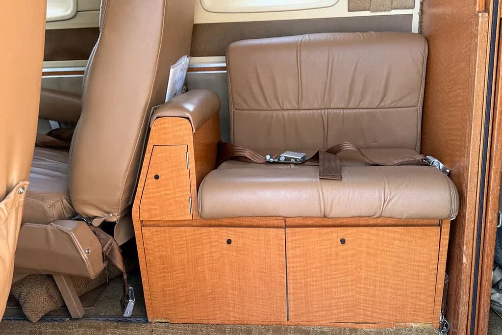 A close-up view of a tan leather passenger seat with a wooden base and storage compartments in the aircraft cabin.
