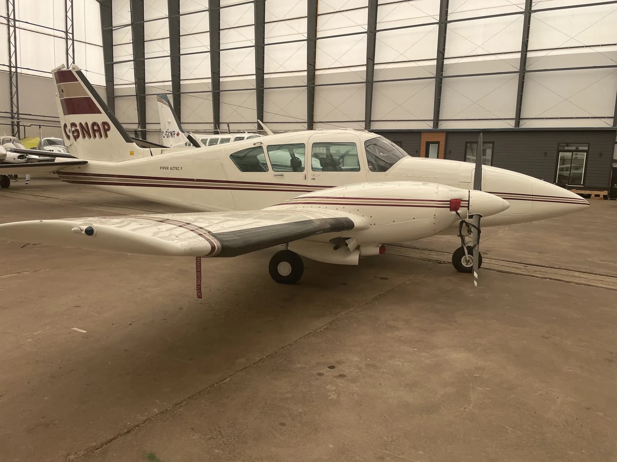 A white Piper Aztec F (C-GNAP) with maroon stripes parked in a hangar, viewed from the front-right.