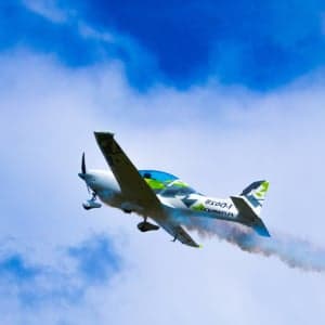 A white and green aircraft flies through the sky, leaving a smoke trail.