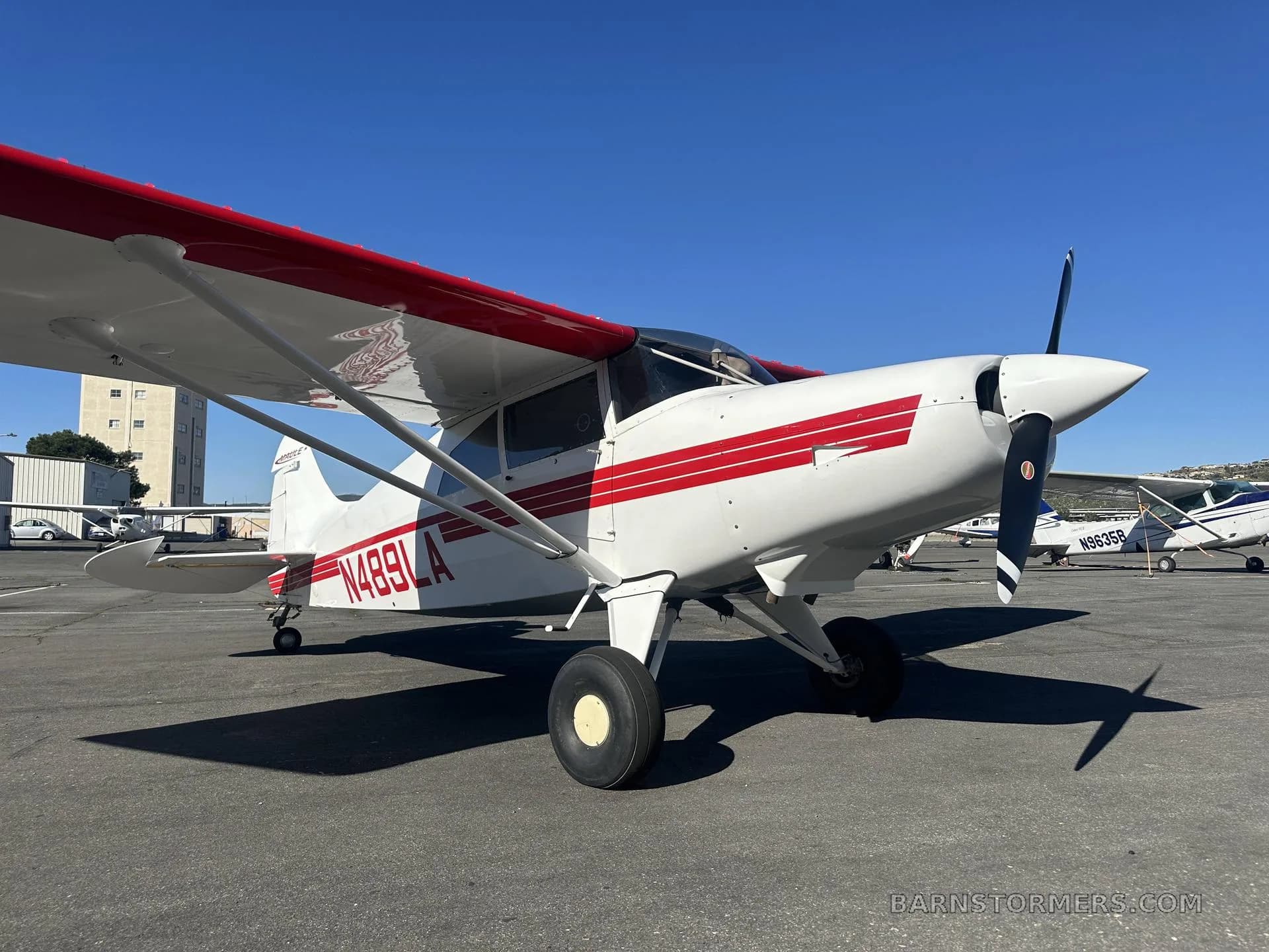 A white Maule M-7-235 aircraft with red stripes and registration N489LA is parked on an asphalt tarmac under a clear blue sky.