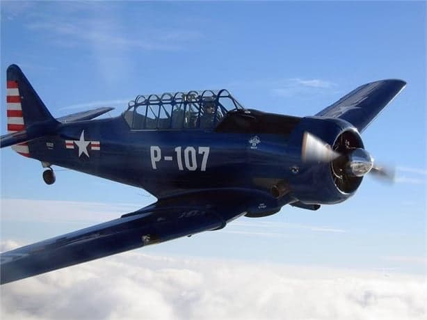 A blue North American Aviation T-6 Texan aircraft in flight with a white star and red and white stripes on the tail.