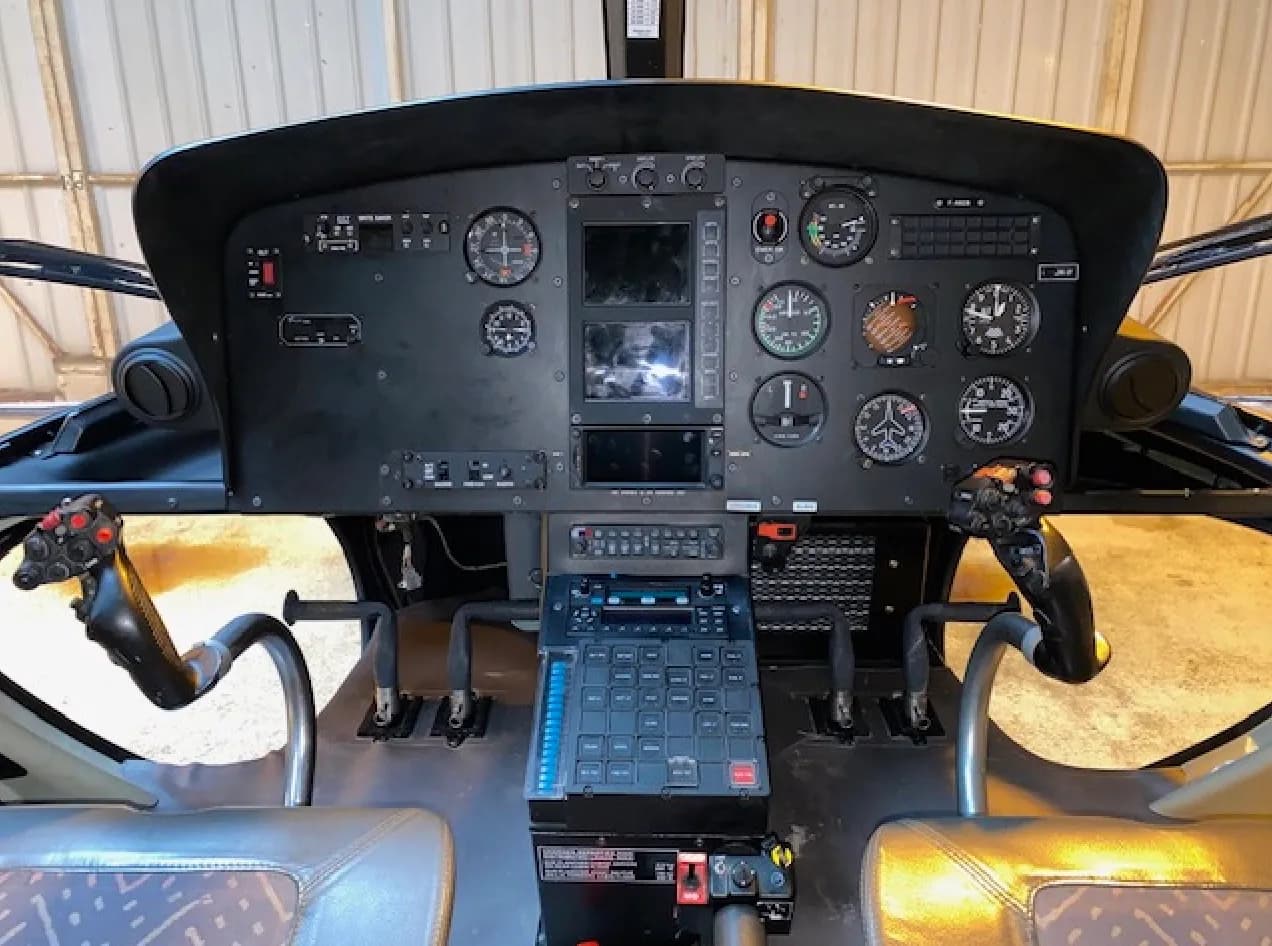 A detailed view of a helicopter cockpit, showing the instrument panel, dual cyclic controls, and collective lever.