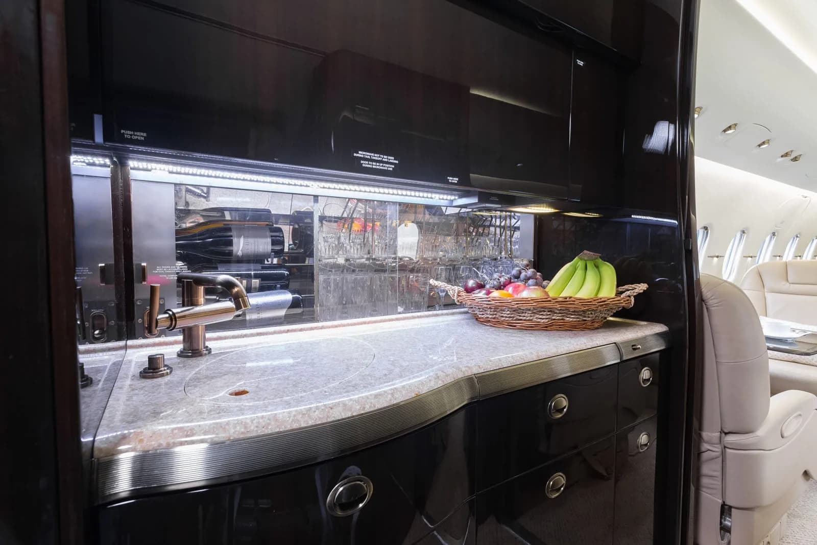 A well-stocked aircraft galley with a sink, dark wood cabinetry, and a basket of fruit, with the cabin visible in the background.