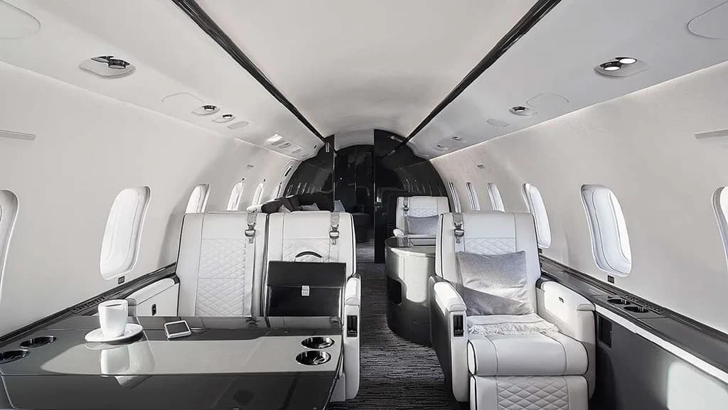 The luxurious interior cabin of a private jet, featuring white quilted leather seats, a dark table, and a black briefcase.