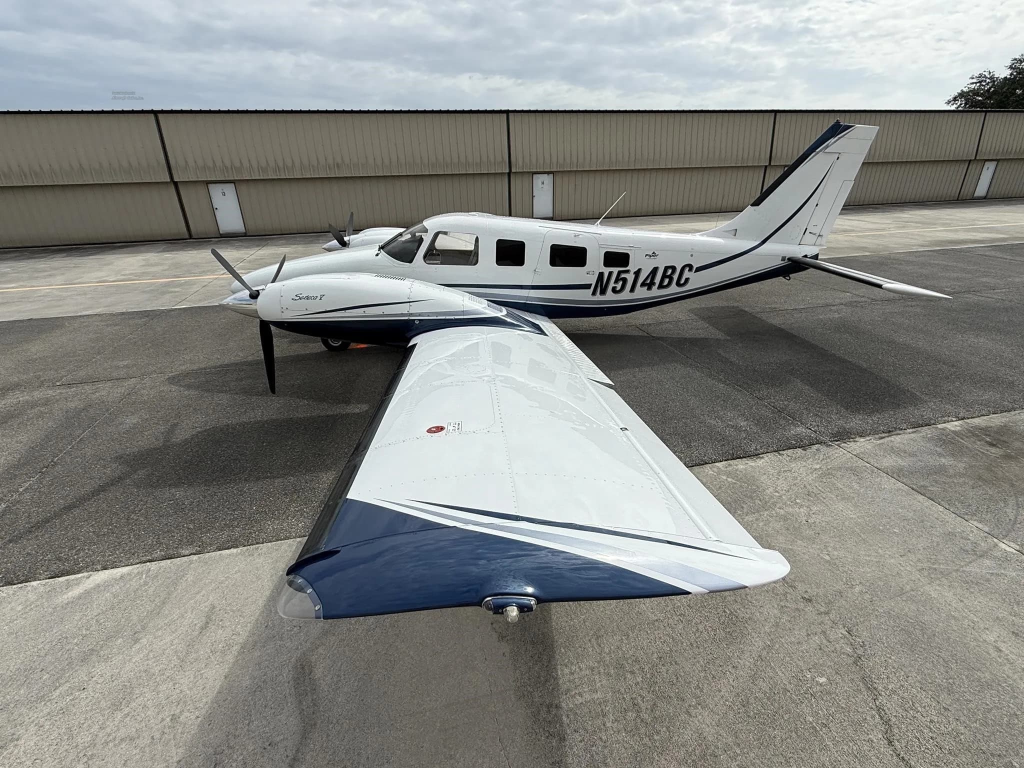 A white and blue Piper Saratoga T aircraft (N514BC) is parked on an asphalt tarmac under a cloudy sky.