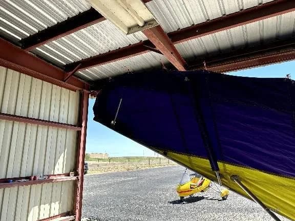 A view from inside a hangar showing the purple and yellow wing of an aircraft, with a yellow ultralight trike on the tarmac outside.