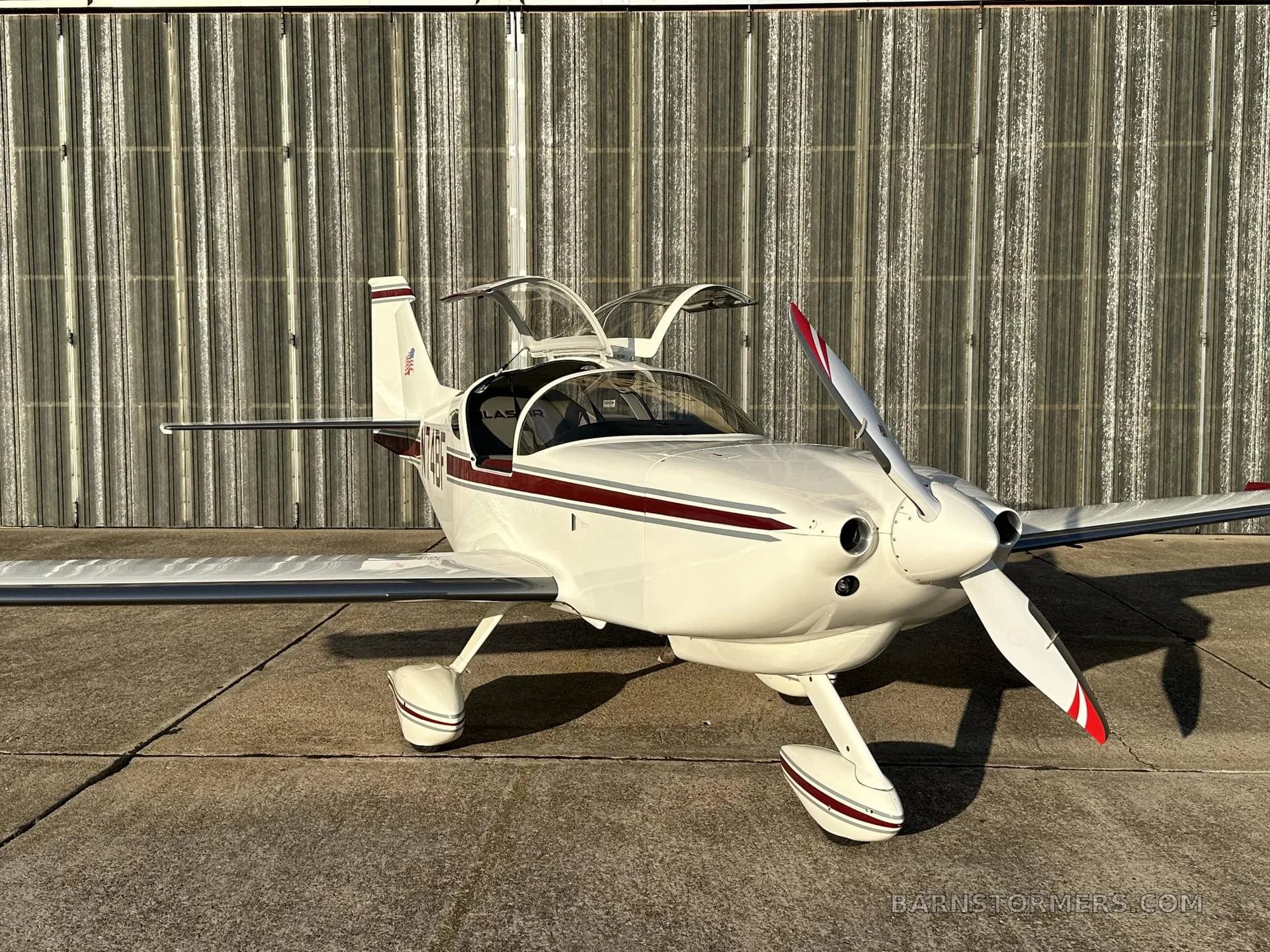A white single-engine aircraft with a maroon stripe and registration N74BF is parked on concrete with its canopy open, viewed from the front-right.